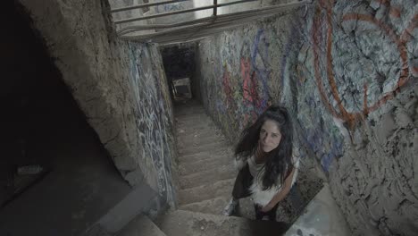 A-young-woman-with-a-staring-look-standing-at-the-top-of-the-stairs,-in-a-narrow-corridor-covered-with-graffiti-with-bars-on-the-ceiling