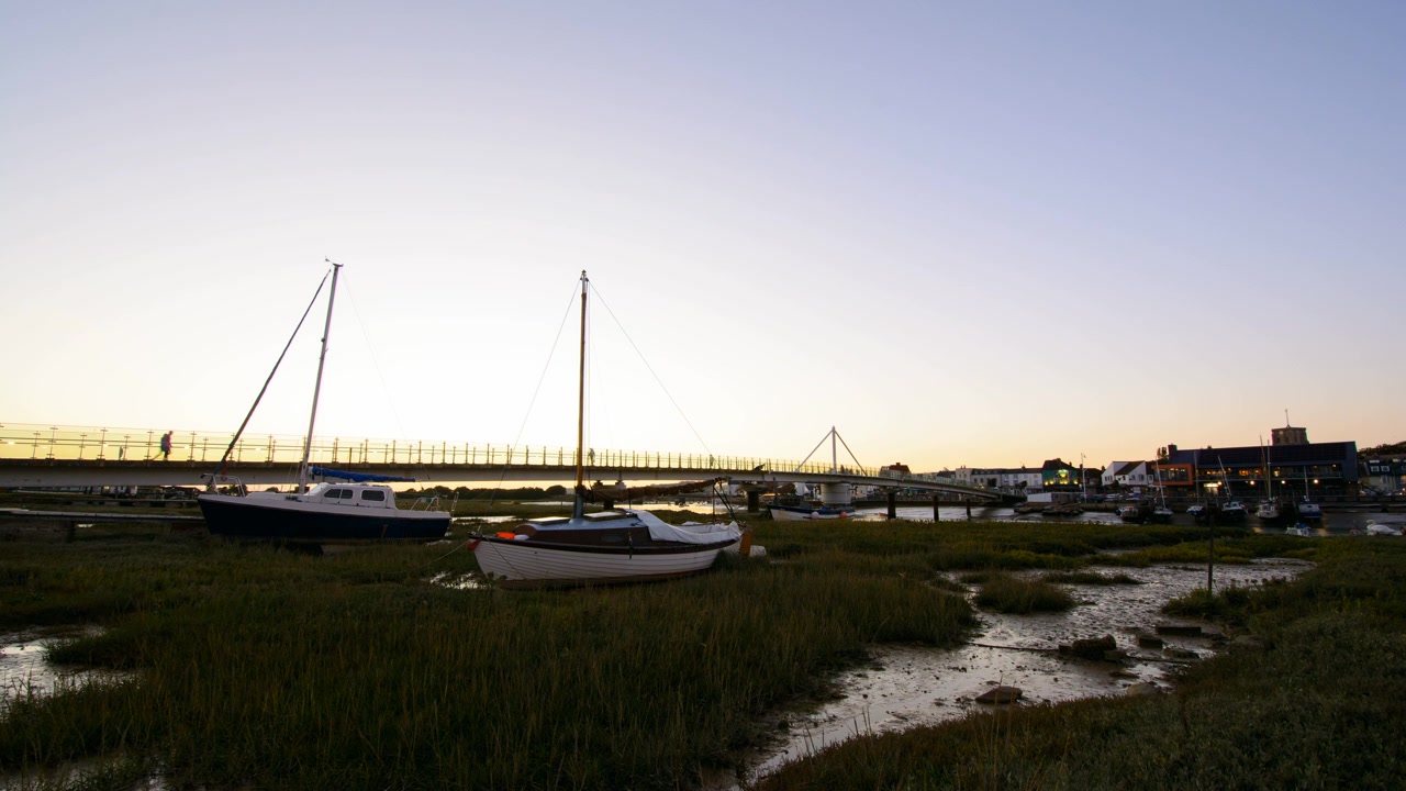 Sunset over a coastal town with boats in a river, transitioning from golden hour to darkness as town buildings and street lights gradually illuminate