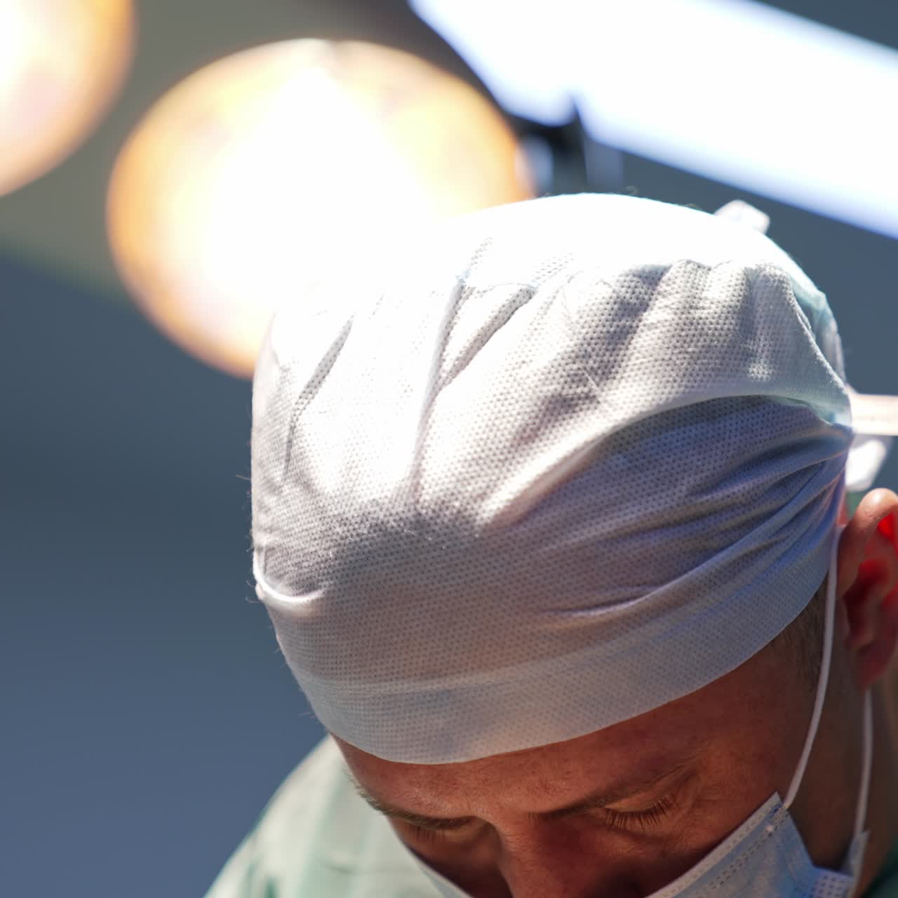 Adult surgeon in mask and cap performing operation. Close up. Lamps in surgery room turned on at blurred backdrop