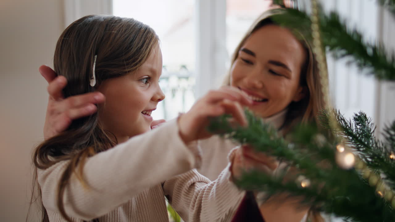 귀여운 아이가 엄마와 함께 크리스마스 트리를 장식하고 있습니다. 엄마가 딸을 포옹하고 있습니다.