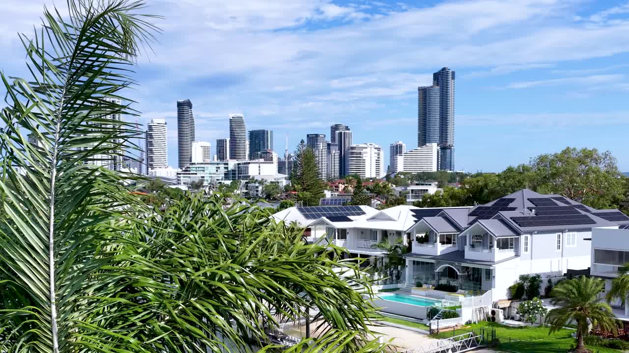 Daytime aerial pan reveals modern canal homes, palm trees, city skyline, and clear blue sky
