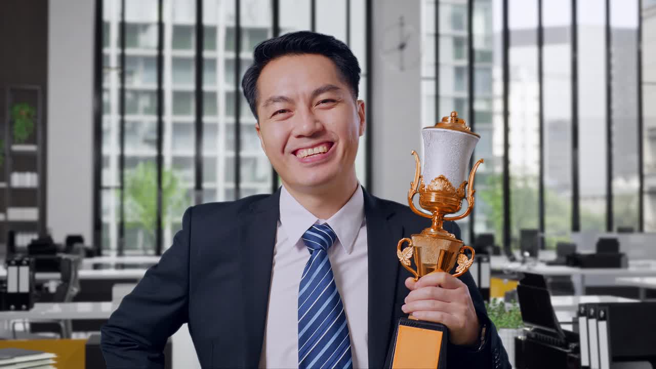 Close Up Of Asian Business Man In A Suit And Tie With A Gold Medal And Trophy Smiling To Camera With One Arm Akimbo Posing As The First Winner In Office