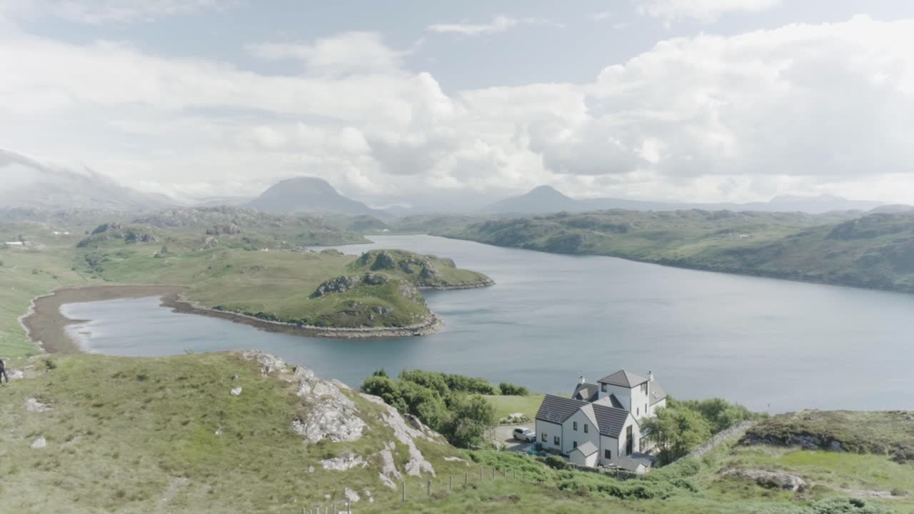 Slow drone panning shot of a house situated on a hill alongside a loch in scotland and tall mountains and clouds in the distance and sun shining