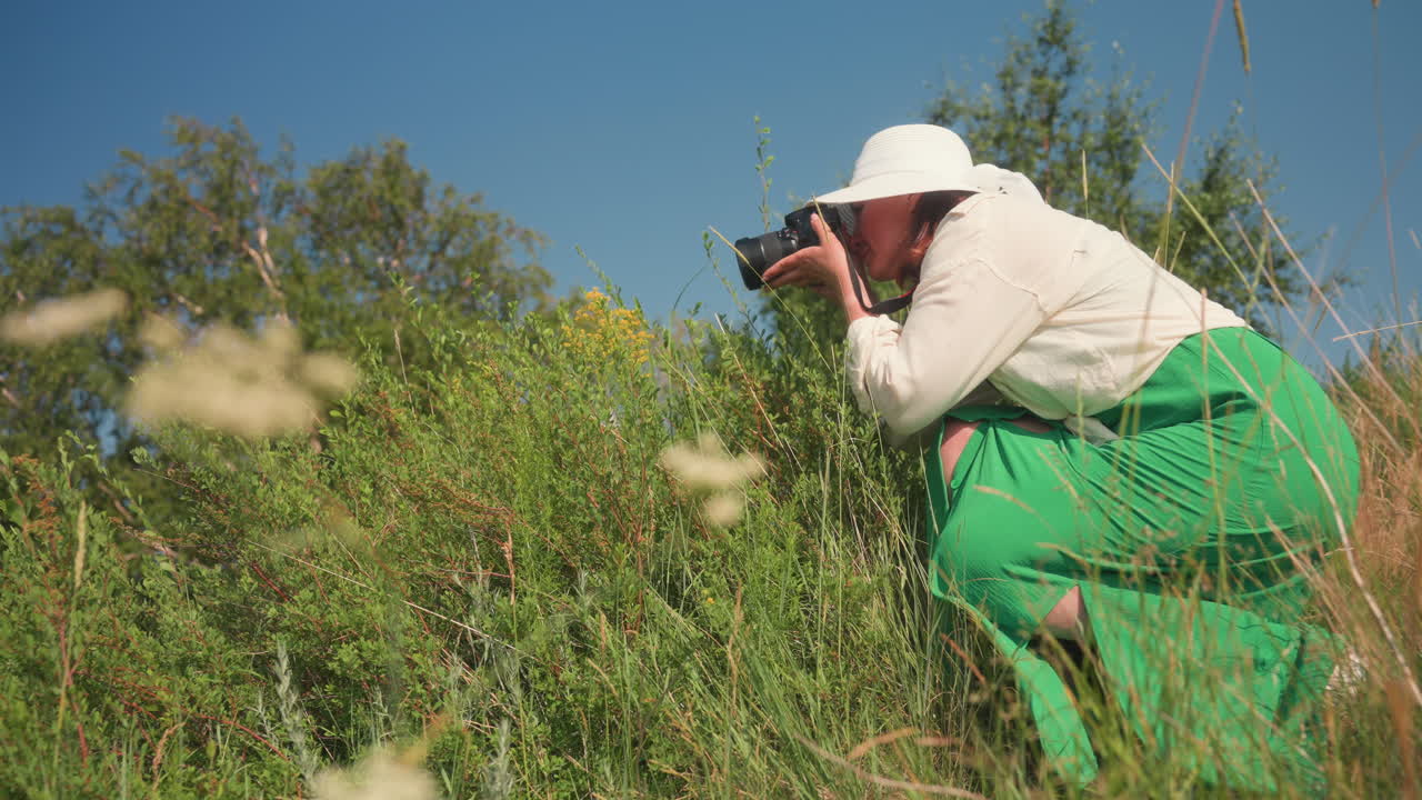 Close up of delicate white wildflower in sharp focus with blurred background showing crouching adult in white hat and green dress taking photo in lush field under clear blue sky on sunny day