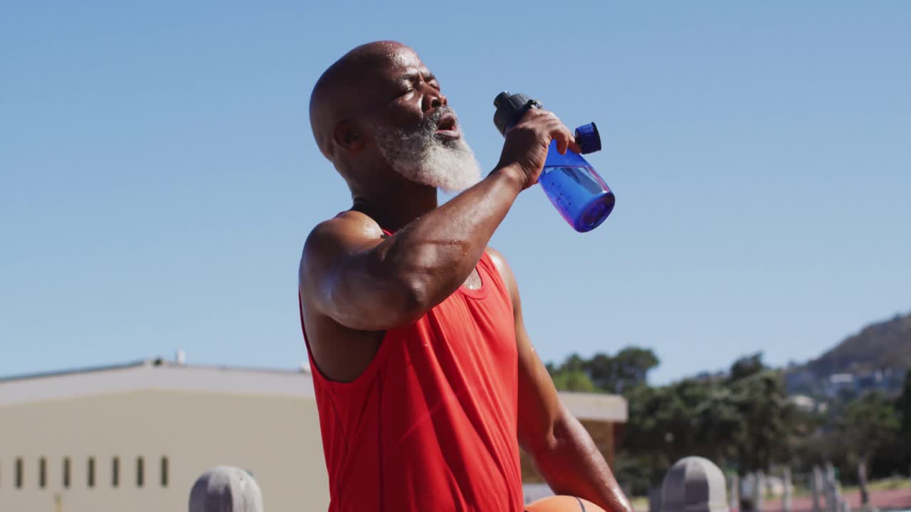 hombre afroamericano sénior con baloncesto bebiendo agua en la cancha cerca de la playa