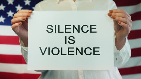 Woman-Holds-Poster-Stating-Silence-Is-Violence-On-American-Flag-Background