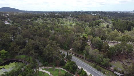 Drone-Volando-Hacia-Un-Gran-Cementerio-Sobre-Un-Jardín-Botánico-Con-Estanque-Y-Cruzando-Una-Carretera