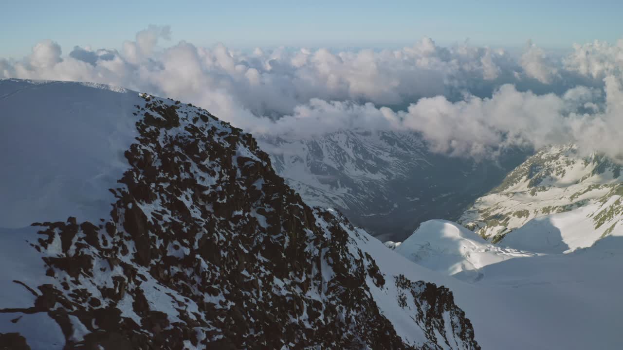 Aerial panorama above snow-capped mountain top, clouds under the summit