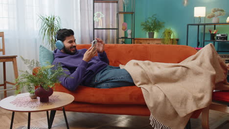 Happy-indian-man-in-wireless-headphones-relaxing-sitting-on-sofa-at-home-listening-favorite-music