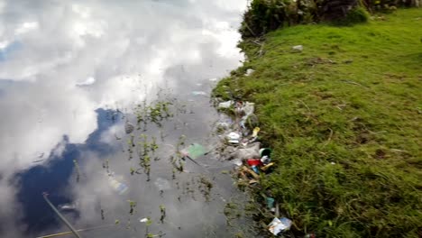 Pan from wooden jetty to clouds reflecting on water to reveal plastic pollution on grassy shoreline