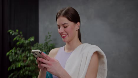 zoom de cerca de una chica morena feliz y alegre en una camiseta púrpura con una toalla blanca en el hombro navegando por las redes sociales usando su teléfono inteligente blanco después de jugar deportes en un apartamento moderno por la noche