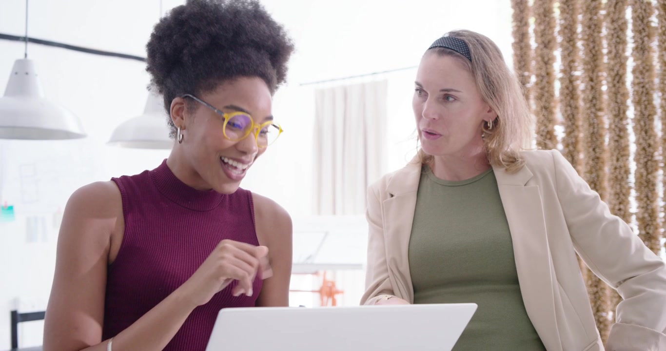 Colleghe donne di diverse etnie che iniziano una dimostrazione in ufficio, con la presentatrice che indica un laptop bianco per la revisione