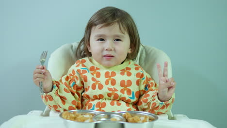 happy 3-year-old girl enjoy eating tasty crispy chicken breast sitting in kid's chair at home, she smiles and shows v gesture