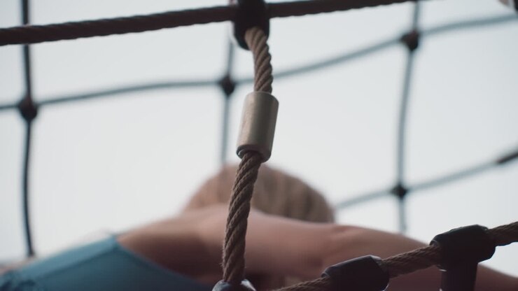 young girl climbing rope net structure in park playground at sunset, showcasing determination, balance and adventurous spirit as she navigates climbing frame under soft evening sky