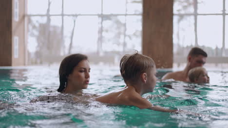 happy family is swimming together in large pool of wellness or hotel parents and children are bathing in warm water