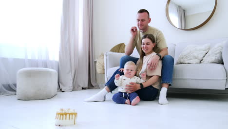 happy caucasian family is at home. parents are watching their baby staring at the cake standing on the floor in distance.