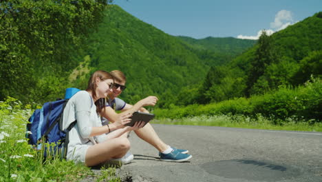 un par de viajeros con mochilas se sientan al costado de la carretera descansan usan una tableta digital