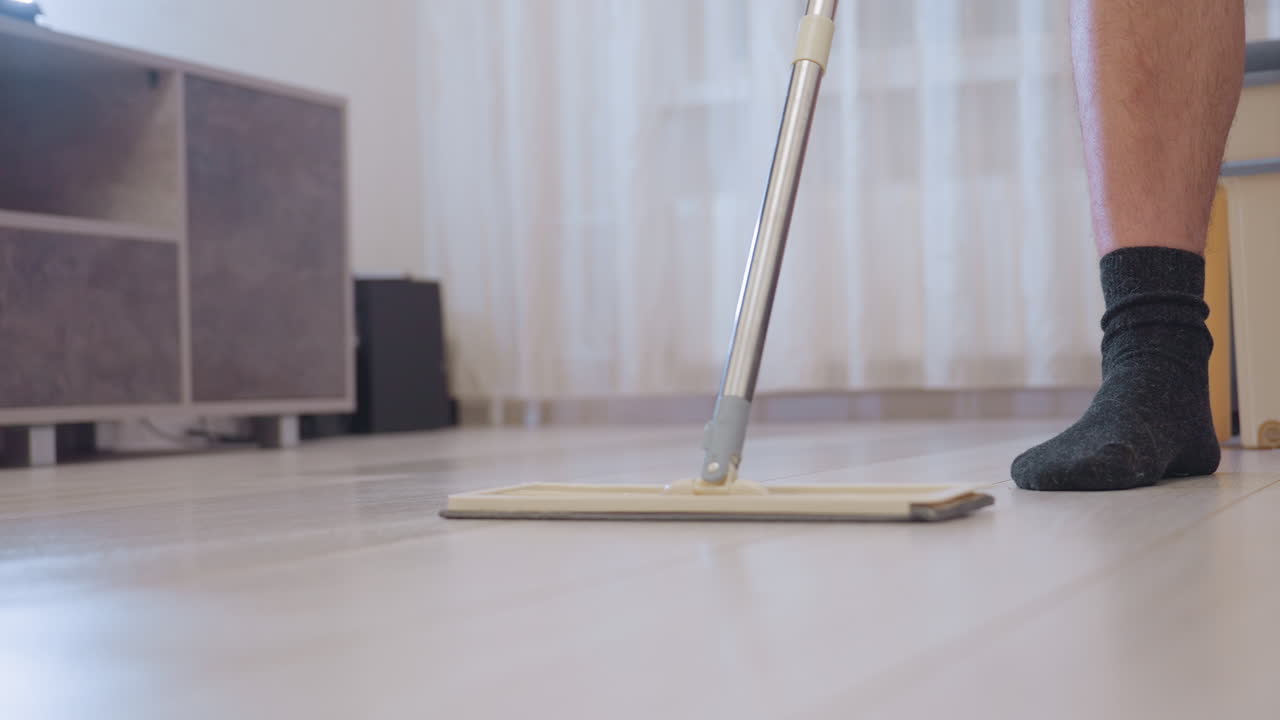 Leg view of cleaner wearing dark socks pushing mop across wooden floor indoors near furniture showing household cleaning routine with focus on hygiene