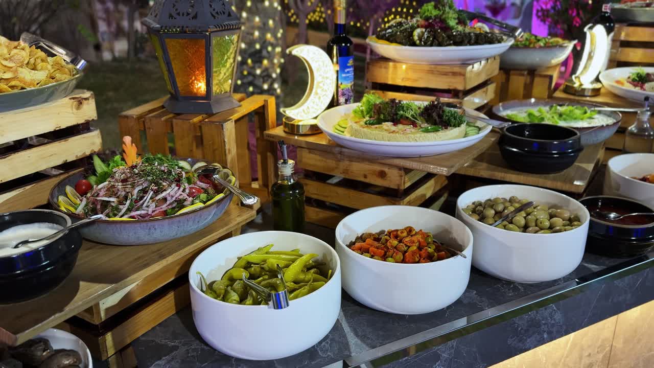 Buffet serale in giardino con ciotole di mezze assortite, insalate, foglie di vite ripiene, olive e salse, esposti su casse di legno con illuminazione a lanterna e a mezzaluna.