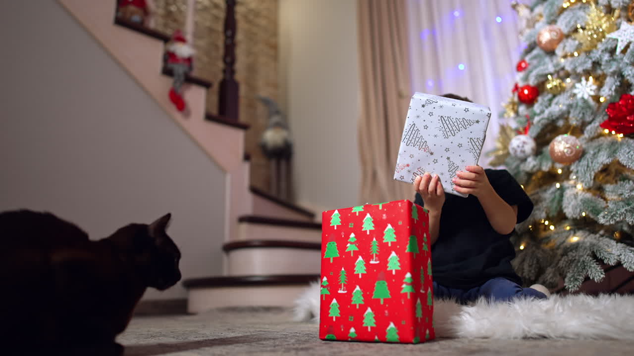 Little kid sitting on the floor near the Christmas tree. Adorable baby boy playing with gift boxes. Black cat watches the child.