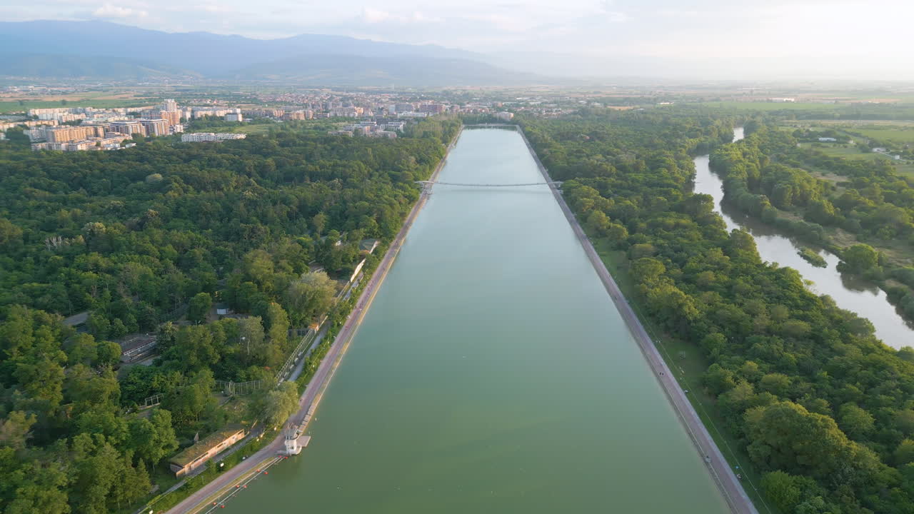 Aerial footage of the Rowing Canal in Plovdiv, Bulgaria, with the Maritsa River to the right, shot during a spring sunset. The lush greenery, soft clouds, and the distant Rhodope Mountains