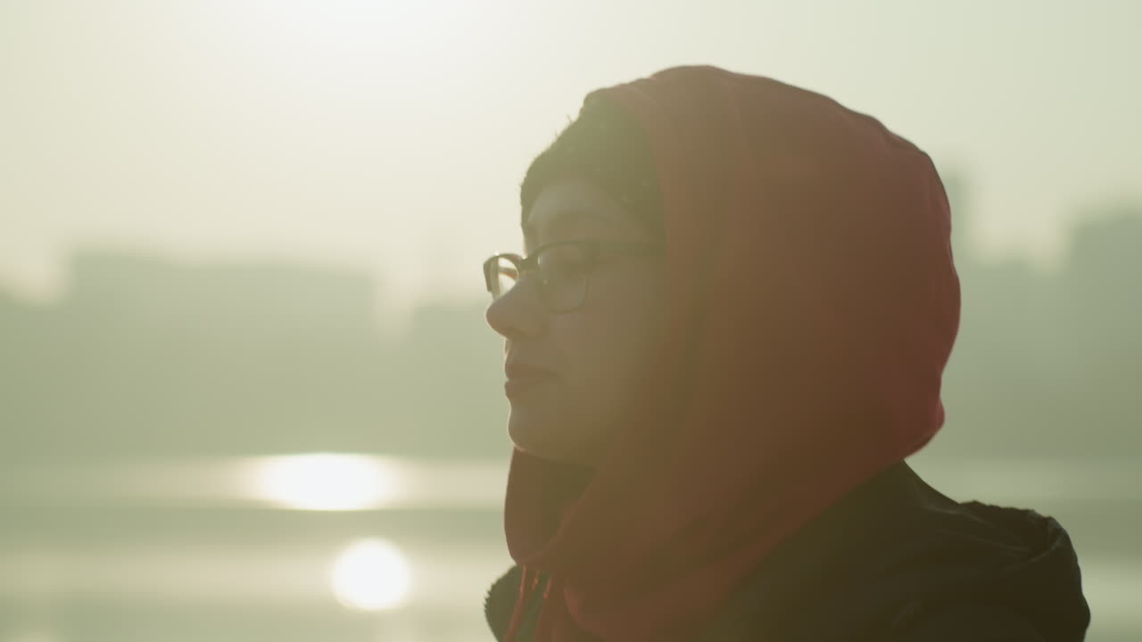 Serene dawn reflection. Asian woman gazes at water. Calm lady with glasses looks over misty cityscape. Peaceful Asian woman in hood watches glowing river at dawn with reflective expression