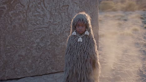 young child in traditional attire made from natural fibers, standing solemnly before an ancient monolith in a sun drenched desert, embodying a timeless connection to history and spirituality