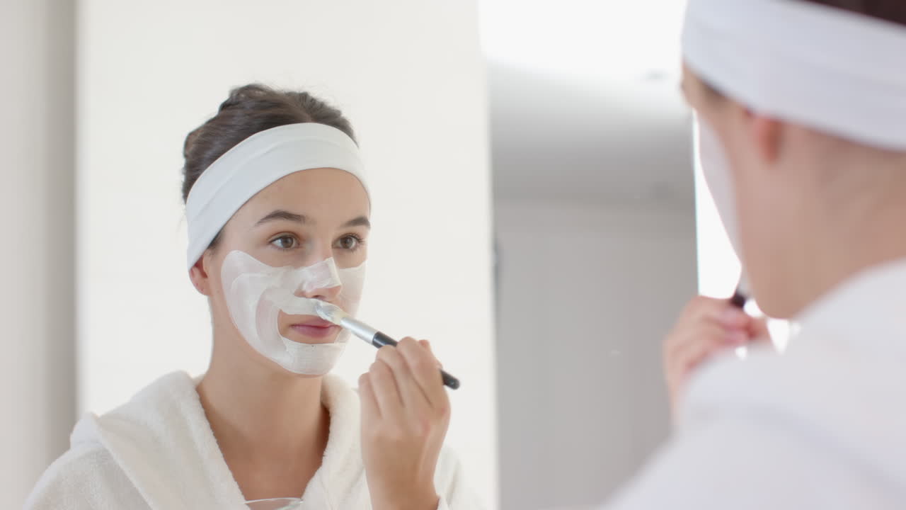 Applying facial mask, woman in bathrobe and headband looking in mirror, at home