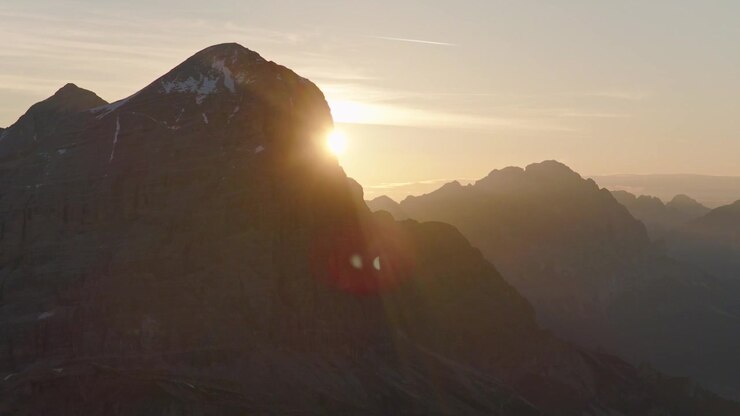 Riprese cinematografiche con drone del sole nascente che emerge dietro una scoscesa cima montuosa delle Dolomiti in Italia. Luce dell'ora d'oro, drammatico paesaggio alpino e scenario aereo mozzafiato