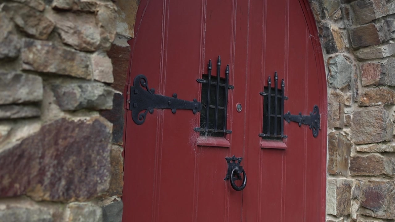 Primo piano suggestivo di una porta di chiesa ad arco rossa e invecchiata con elementi in ferro battuto, incastonata in ruvide mura di pietra. La luce soffusa del giorno illumina le texture, evocando un fascino vintage e un'atmosfera storica.