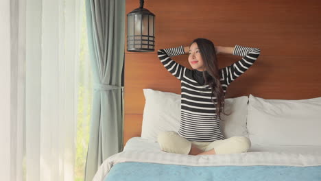 Asian-woman-sitting-on-a-bed-stretching-her-arms-overhead-and-putting-them-behind-her-head-then-leaning-on-a-wall-looking-relaxed-and-happy