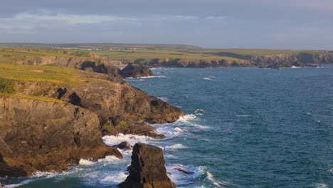 Waves-crash-into-jagged-rocky-seashore-of-Cornish-coastline-in-UK,-aerial