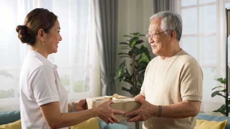 a helper hands a carefully wrapped package to an elderly man in a bright, welcoming living room. the atmosphere is warm and friendly, showcasing a thoughtful interaction between them.