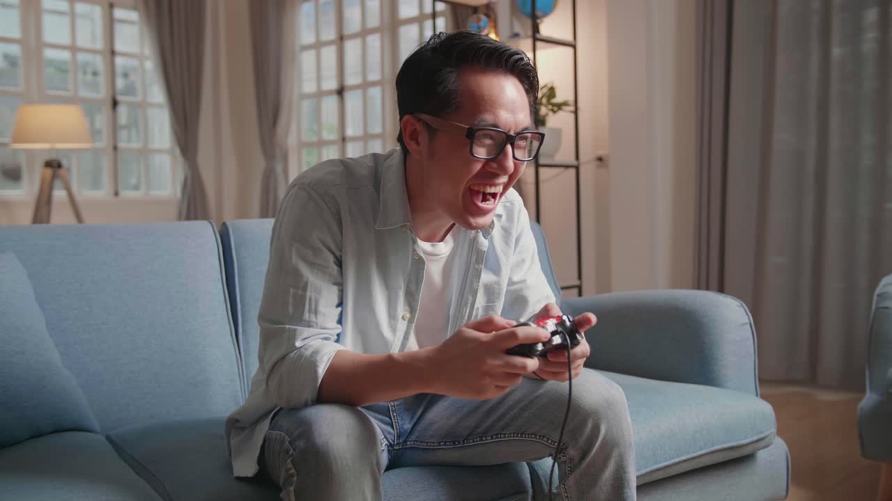 Excited Man Wearing Sunglasses Sitting On A Couch In Living Room, Laughing While Playing Game