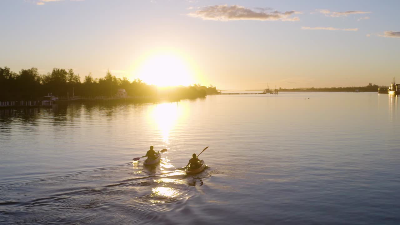 hermosa toma de seguimiento de dos kayaks navegando a lo largo de la costa australiana hacia la puesta de sol, con árboles y cielo azul en el fondo