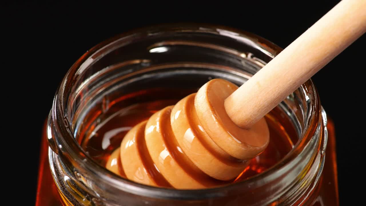 Honey Dripper Is Dipped In A Jar Full Of Delicious Sticky Honey. Healthy Organic Thick Honey On The Black Background, Sweet Honey Dessert 4K. Close Up, Slow Motion