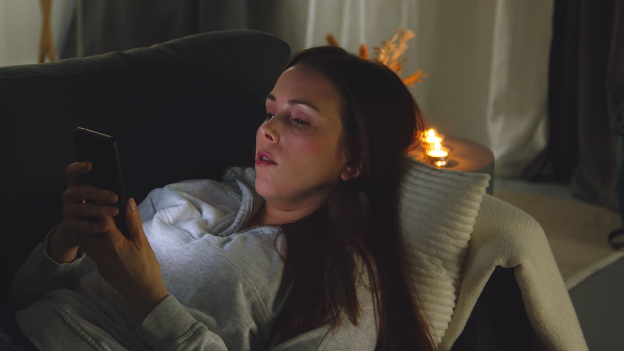 женщина лежит на диване дома по ночам, просматривает онлайн-контент на мобильном телефоне