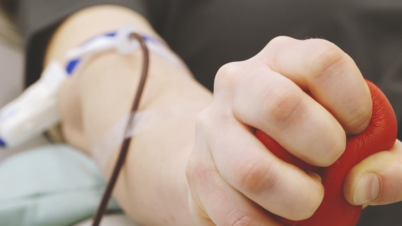 Primo piano di un donatore di sangue sdraiato su una sedia clinica con ago e tubi attaccati, che stringe una palla antistress rossa durante il processo di donazione del sangue in un ambiente medico