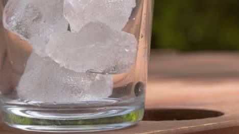 close up of textured ice cubes falling into clear clean glass on picnic table during summer - 4k slow motion footage