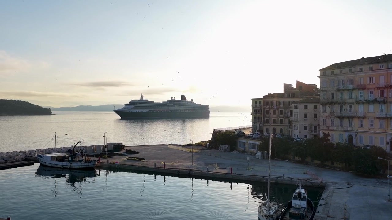 Una vista pittoresca di un'enorme nave da crociera di lusso in partenza dal porto di Corfù, Grecia, sotto il sole del primo mattino. L'architettura costiera classica in stile veneziano, le auto parcheggiate e un porto calmo completano la scena.
