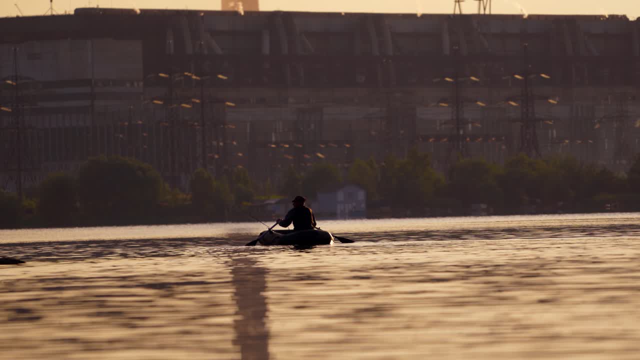 Old man in inflatable boat sailing in the evening. Fisherman in a rowboat with two oars on the background of a big plant. Man tries to find a good place for fishing in the river.