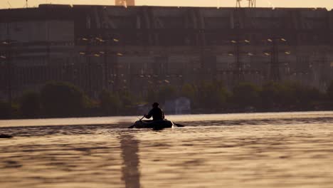 old man in inflatable boat sailing in the evening. fisherman in a rowboat with two oars on the background of a big plant. man tries to find a good place for fishing in the river.