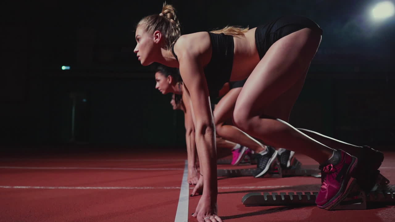 Female runners at athletics track crouching at the starting blocks before a race. In slow motion.