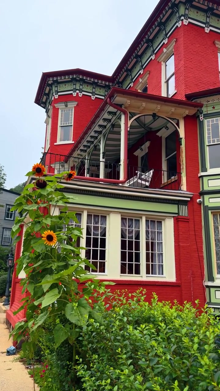 Maison victorienne en briques rouges avec des boiseries vertes et des tournesols. Extérieur d'une maison victorienne en briques rouge vif, dotée de boiseries décoratives vertes, d'un balcon et de grands tournesols en fleurs.