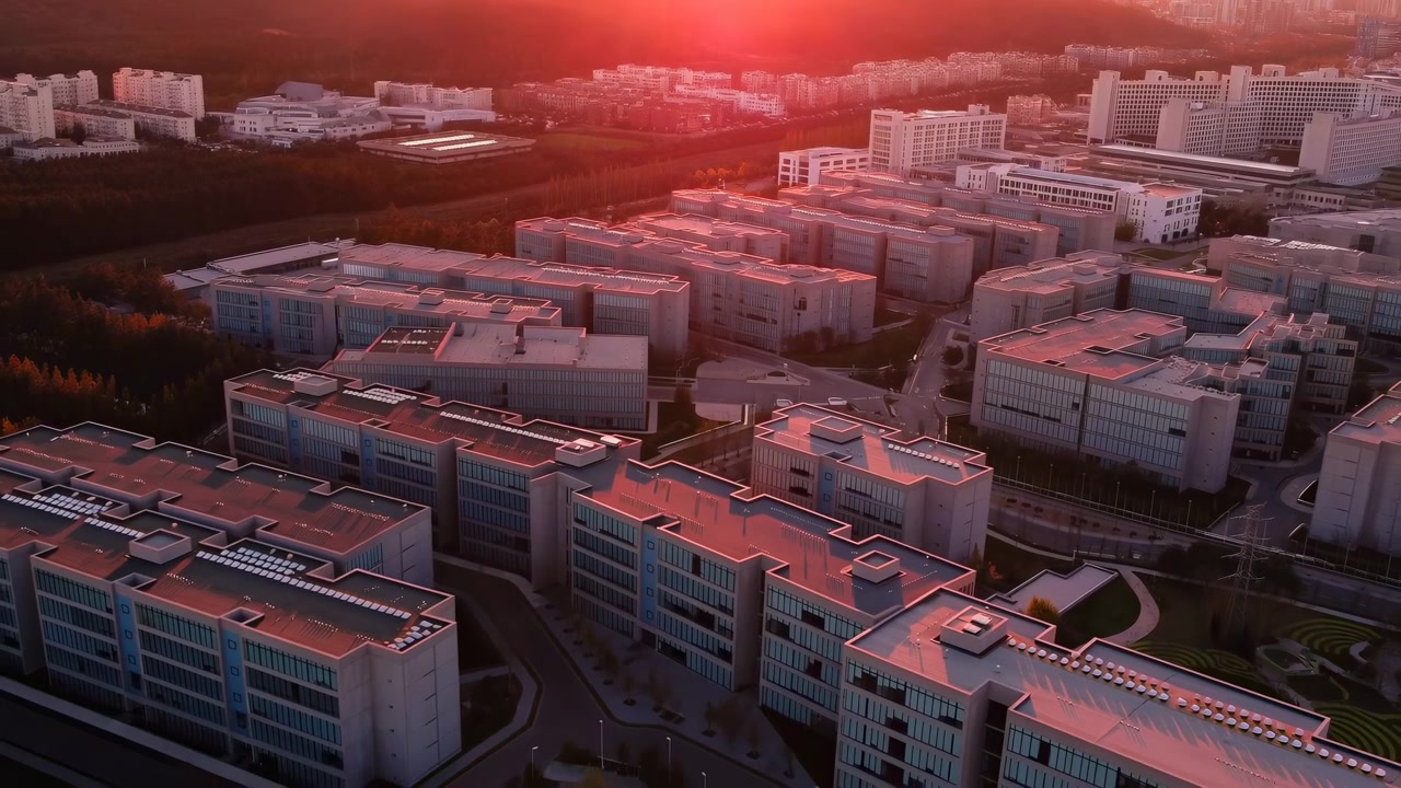 Letecký záběr firemního kampusu při západu slunce, střechy září červeným světlem, moderní budovy a klikaté silnice, dron.