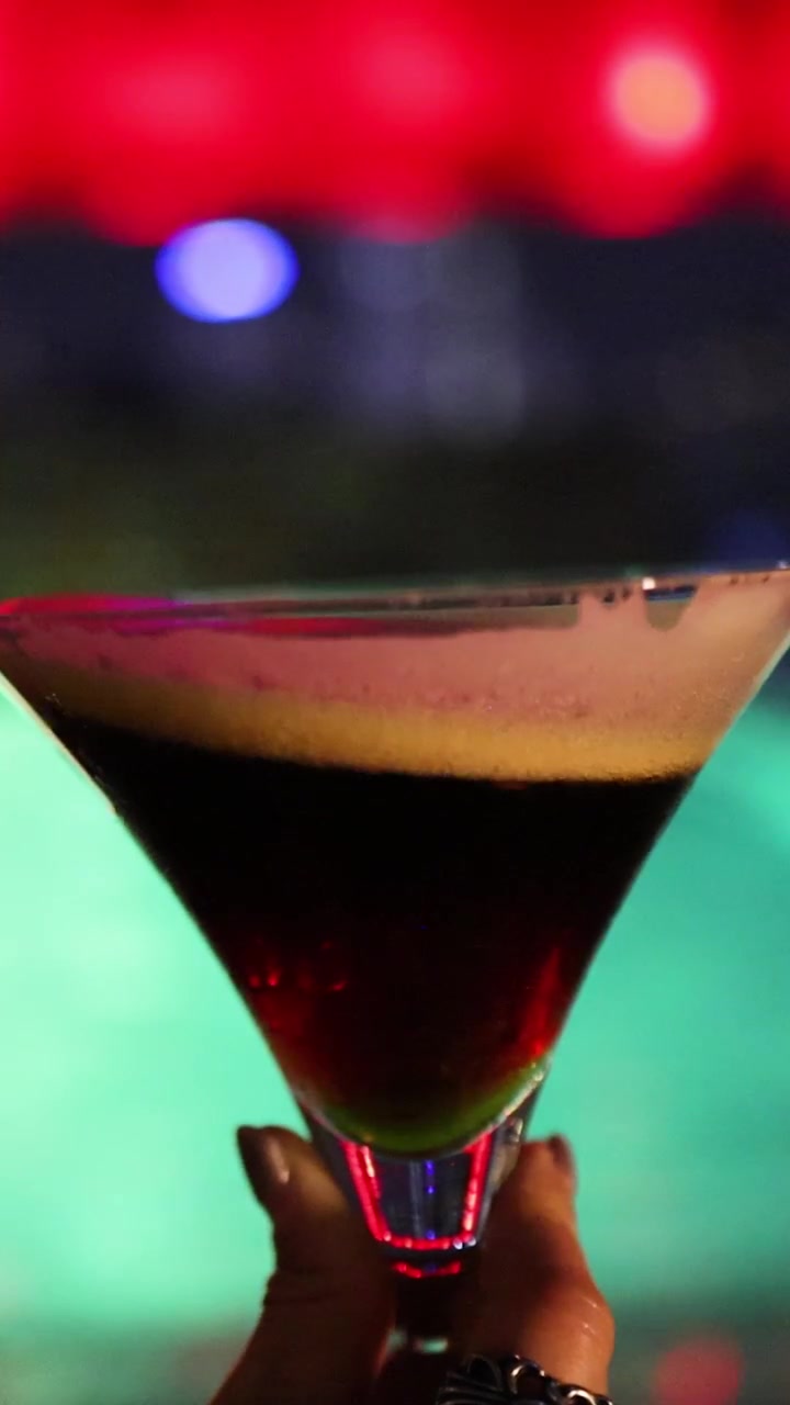 Una mano mescola delicatamente un espresso martini in un bicchiere da martini, sullo sfondo di luci colorate e sfocate di un night club con un'illuminazione suggestiva e soffusa.