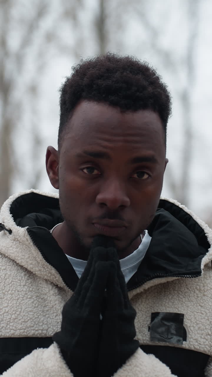 Young boy in black gloves and white winter jacket standing outdoors during winter, hands pressed together in deep thought, with a blurred background of bare trees and snow