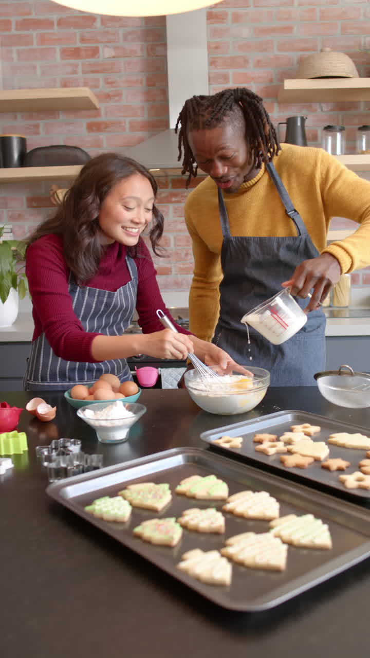 Vertical video of happy diverse couple in aprons baking christmas cookies at home, slow motion