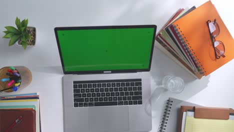 vista de arriba hacia abajo de una computadora portátil con pantalla verde simulada pantalla chromakey en un escritorio de oficina de madera al lado de la libreta con bolígrafos, gafas y un vaso de agua.