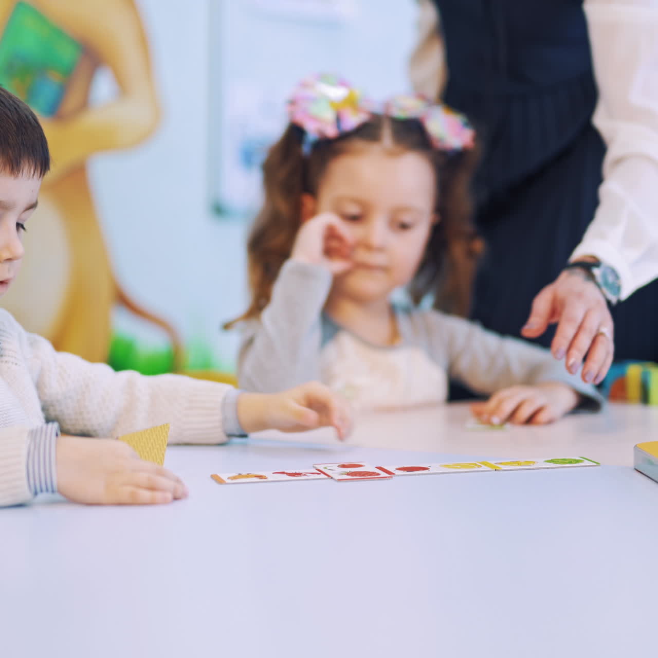 Little pupils are studying at elementary school. Teacher helps children to put picture cards into the necessary place on the table during the lesson.